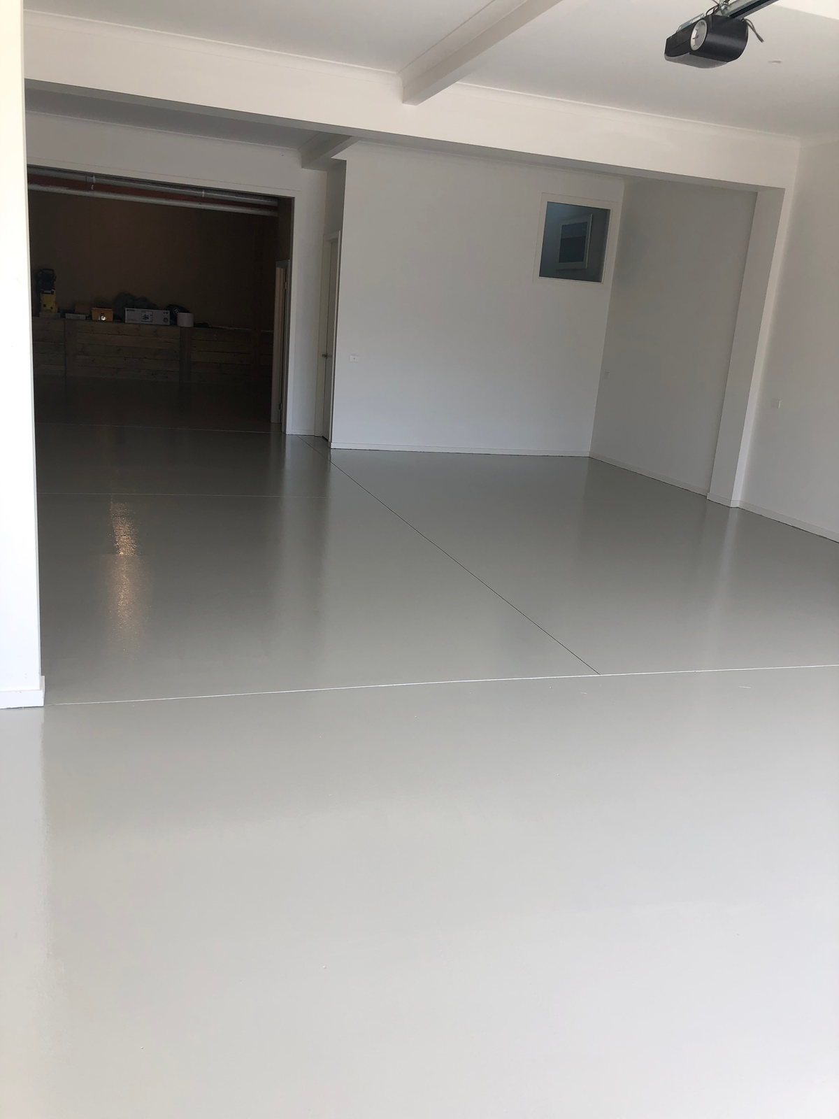 The same garage floor after coating: glossy grey epoxy reflecting ceiling light, with a roller door at the far end