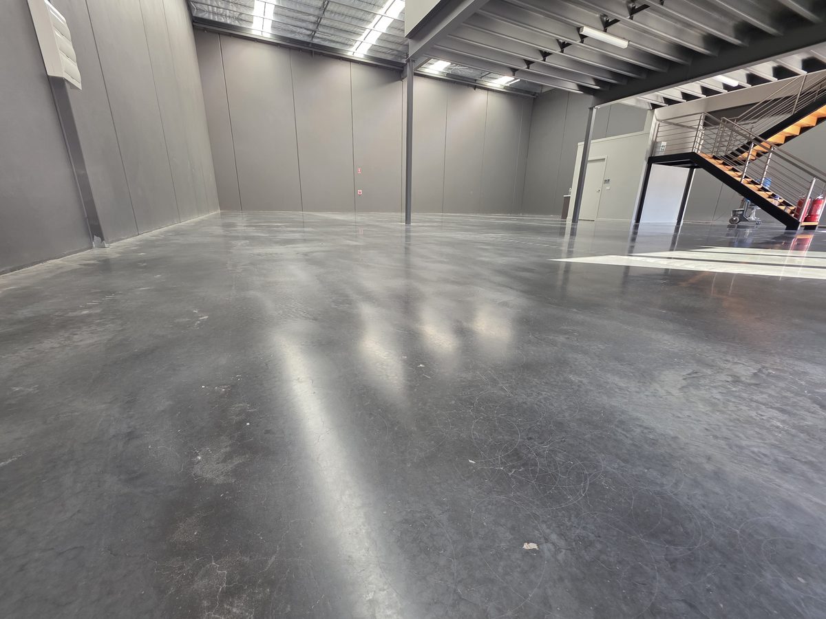 Factory-spec polished concrete floor in an empty industrial unit, showing a soft even sheen and visible saw-cut joints beneath a steel mezzanine