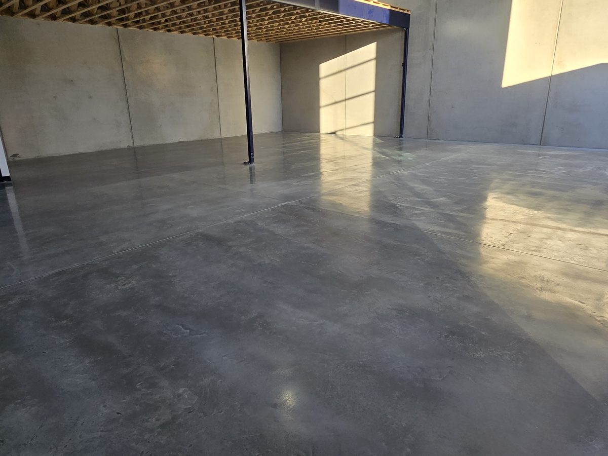 Factory-spec polished concrete floor in a precast-panel warehouse, with afternoon sunlight raking across the surface below a timber-joist mezzanine