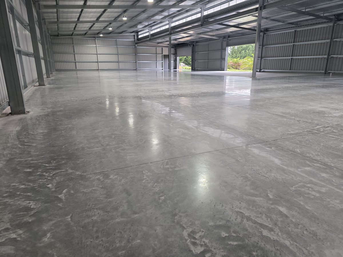 Finished factory-spec polished concrete floor running the length of an empty steel-framed warehouse bay, with overhead lights reflecting evenly across the slab