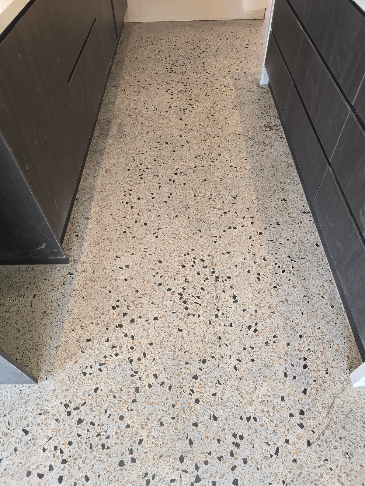 Pale grind-and-seal floor with dark exposed aggregate running between dark kitchen cabinetry