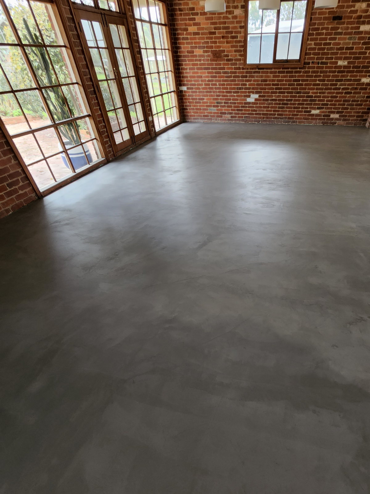 Seamless grey microcement floor in a room with exposed red brick walls and timber French doors